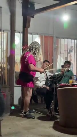 Here is a description of the video:

An adult person in a pink dress and blonde wig is playing the drums for two seated people at an outdoor gathering. The person is using drumsticks and seems to be entertaining the two men. One of the men is smiling and appears to be enjoying the performance, while the other is holding a smartphone and recording. The setting seems to be a backyard or patio with concrete flooring and some foliage. A partially visible table with glasses and a bottle indicates a casual social event. The space is illuminated by overhead lights, and a curtain or drape can be seen in the background through glass doors.