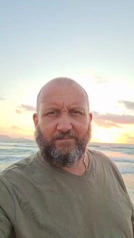 A man with a bald head and a full beard is standing on a beach. He is wearing a green t-shirt and a gold chain. The ocean is visible behind him, with waves rolling in. The sky is a pale blue with hints of orange and yellow from the setting sun. The man is looking directly at the camera with a serious expression.