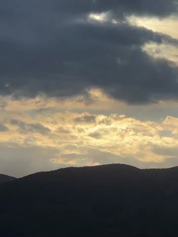 The video shows a silhouette of a mountain range against a cloudy sky. The clouds are dark gray in the upper part of the frame, and lighter, yellowish-white closer to the horizon. Some rays of sunlight peek through the clouds, creating a dramatic effect. The wind sounds are audible.