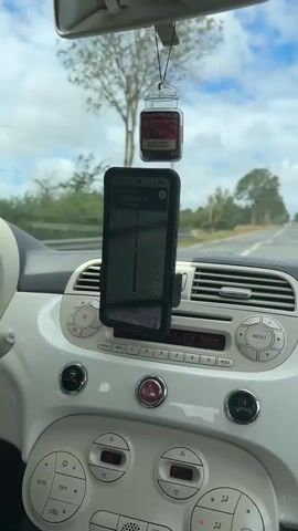 The video shows the interior of a car with a smartphone mounted on the dashboard, displaying a navigation app. The phone is positioned vertically in a black holder. The navigation app shows a route with upcoming turns and road names, such as "Rue du Château" and "D901". A car air freshener in a small jar with a dark liquid and a label reading "PINK zu CANDLE" is hanging from the rearview mirror. The car's radio is visible below the phone mount, with buttons and a display showing "191.50 MHZ" and other labels. The dashboard is white. Outside the car, trees and a cloudy sky are visible through the windshield. The time on the phone changes from 12:00 to 13:00.