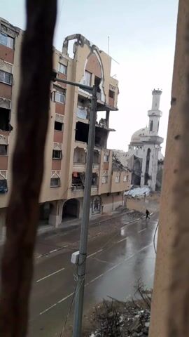 From behind a damaged structure, we see a street with substantial damage to the buildings around it. On the left, a multi-story tan building has large sections missing and is pockmarked with holes. Laundry is hanging from the damaged structure. A tall light post stands in front of the building. Across the street, a mosque is also heavily damaged with its dome and tower partially destroyed. A single person walks down the wet street. Debris is scattered on the ground. The sky is overcast.