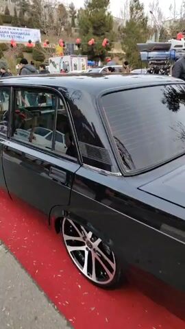 The video shows two cars on display at a car show. The first car is a black Lada with large, silver rims. The second car is a dark blue BMW with silver rims. Both cars are parked on a red carpet. In the background, there are people walking around and looking at the cars. There are also red and black balloons hanging above the area. A banner for the "II XALQARO NAMANGAN AVTO FESTIVAL" is visible in the background.