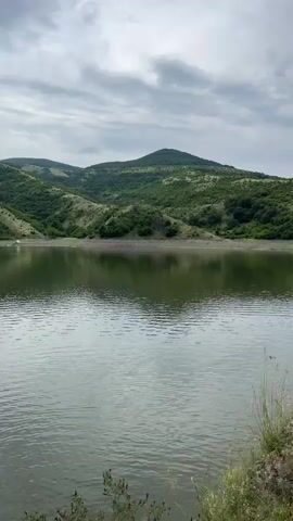 The video features a view of a tranquil lake surrounded by lush green hills. A long suspension bridge extends across the water, its reflection visible on the surface. The sky is overcast with a mix of white and gray clouds. A few small birds can be seen flying low over the water. The camera slowly pans to the right, revealing more of the bridge and the surrounding landscape. The water's surface is slightly rippled, reflecting the overcast sky.