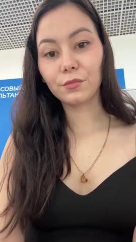 The video shows a close-up of a young woman with long dark hair and a black tank top. She is wearing a necklace with a heart-shaped pendant. She has makeup on and is looking directly at the camera. In the background, there is a blue sign with white text, partially visible. The ceiling has a grid pattern. The woman briefly touches her hair and then looks back at the camera.