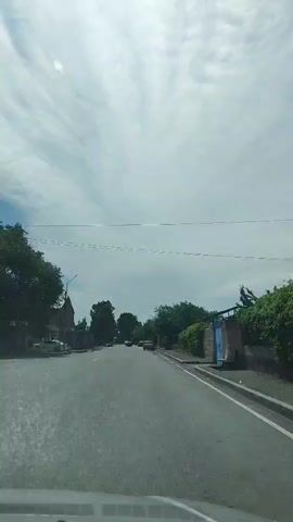 The video shows a view from inside a vehicle traveling down a paved road. The sky is cloudy with streaks of white clouds. On the left side of the road, there are buildings and trees. On the right side, there is a wall covered in green foliage and more buildings. Several cars are visible on the road, and a person can be seen walking in the distance. The overall impression is that of a suburban or rural road on a cloudy day.