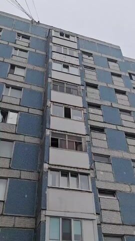 The video shows a close-up, downward-angled shot of a tall apartment building. The building's facade is covered in a pattern of blue tiles and grey rectangular panels. A central section of the building features a vertical arrangement of balconies, some of which are enclosed with windows, while others are open. Several windows on the building are open or appear to be damaged. The lighting suggests it is daytime, and the sky is overcast. The camera movement is primarily downward, with slight swaying. No people or other significant objects are visible outside the building itself.
