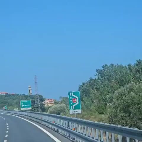A video captures a sunny day with a clear blue sky. The perspective appears to be from a vehicle on a highway. On the right side of the frame, a green highway sign with white text indicates an "SOS" emergency call box and states "250 m". Further down the road, another green sign points left towards "GENOVA" and right towards "ALBENGA", with "1500 m" indicated below. In the background, a tall electrical tower stands prominently, with a church steeple visible to its left, peeking above lush green trees and some buildings. The overall scene is one of a rural or suburban highway setting under bright daylight.