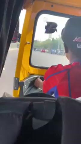 The video shows the interior of a yellow auto-rickshaw or tuk-tuk while in motion on a city street. The camera is positioned behind the passenger seat, facing forward towards the driver. The driver, wearing a black baseball cap with "Atlanta" embroidered on it and a red and blue shirt, is operating the vehicle. Outside the window, other vehicles, including motorcycles and a red car, can be seen moving along the road. Buildings and trees are visible in the background. The overall environment appears to be a busy urban area. The sound of wind is clearly audible.