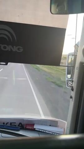 The video provides a first-person perspective from inside a vehicle, likely a bus or truck, as it travels along a paved road. The scene is characterized by a forward-facing view through the windshield, revealing a highway with a white line dividing the lanes. To the right, a parallel road or shoulder is visible, bordered by green vegetation and grass. The sky transitions from a bright, hazy area at the horizon, suggesting late afternoon or early morning light, to a clear blue overhead.

Inside the vehicle, several objects are visible. A dark sun visor with a logo featuring concentric arcs and the text "ONG" is prominent in the upper portion of the frame. Below this, on the dashboard, are various items including papers, a red and white package that reads "АПТЕЧКА" (meaning "first-aid kit" in Russian), and a blue object that could be a pen or marker. A steering wheel is partially visible in the lower part of the image. A rearview mirror, positioned at the top center, reflects a scene from within the cabin, showing at least two individuals. One person appears to be holding a phone, and another person is visible in the reflection, possibly looking at the camera or out the window. A sticker with red lettering, partially obscured, is affixed to the interior above the windshield.

The overall impression is a journey through a rural or semi-rural landscape, likely in Kazakhstan, given the location context. The time of day appears to be either sunrise or sunset due to the warm light on the horizon. The activity is transportation, with the vehicle in motion.