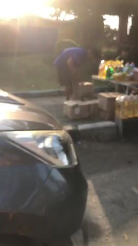 The video shows an outdoor scene at dusk with a bright sun flare obscuring some details. In the foreground, the front of a dark-colored car is visible. To the right of the car, a person wearing a blue shirt and dark shorts is bending down, interacting with several cardboard boxes stacked on the ground. Behind this person, a table is set up with numerous bottles of drinks, some in plastic and others in glass, arranged on it. The ground appears to be dirt or gravel. The lighting suggests it is late afternoon or early evening.