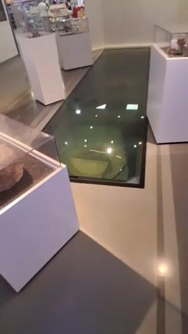 The video shows a person walking on a glass-covered floor. Beneath the glass, there is a display that appears to be a circular object submerged in water. The environment seems to be a museum or exhibition space with white display stands visible.