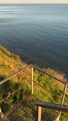 A vertical video captures a serene ocean view from a clifftop at sunrise. The foreground features a rusty metal railing and chain-link fence, with dry grass and green vegetation clinging to the cliffside. Beyond the railing, the vast expanse of the calm sea stretches to the horizon, reflecting the soft hues of the rising sun. The sky transitions from a pale yellow near the horizon to a clear, light blue overhead. Small ripples on the water suggest a gentle breeze.

The sun, a bright orb, is visible on the right side of the frame, casting a shimmering golden path across the water. A few distant boats or buoys can be seen on the horizon. In the lower right corner of the last frame, a person can be seen standing near the railing, seemingly enjoying the view. The overall mood is peaceful and tranquil, evoking a sense of calm and natural beauty. The time of day is clearly sunrise, indicated by the sun's position and the warm, soft light. The location is identified as Tsibanobalka, Russia. There is no visible text in the video.