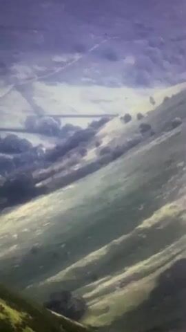 A drone or camera flies over a mountainous landscape. The terrain is covered in green vegetation and dotted with trees. The sky is cloudy with a hint of purple. In the distance, a white object, possibly an airplane or another drone, flies across the screen, leaving a trail of light. The camera pans down and to the right, revealing more of the green hills and valleys below. The video is shaky and the image quality is poor, suggesting it was taken with a handheld device or under low light conditions.