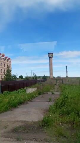 The video captures a view from a moving vehicle, showing a four-story building with a unique architectural style, featuring arched windows on the top floor and a tan brick exterior with pink accents. Several flags are displayed in front of the building. Adjacent to the building, separated by a fence, is a large, grassy area with an older, cylindrical water tower in the background. The sky is bright blue with scattered clouds. The overall scene suggests a transition from a rural or industrial area to a more developed one, with the building possibly serving an administrative or commercial function.