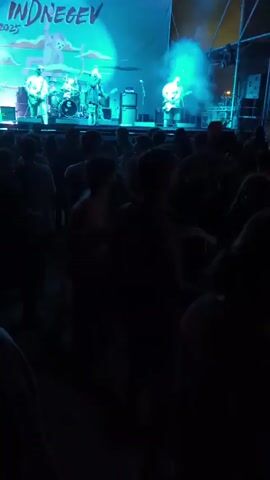 A band performs on a stage under bright blue and white lights in front of a large crowd at night. A large blue banner reading "INDNEGEV 2025" hangs behind the band. The crowd is a dense mass of people, some jumping and raising their arms in the air. Smoke billows from the stage, partially obscuring the band. The band consists of multiple members playing guitars and drums. The setting appears to be an outdoor music festival.
