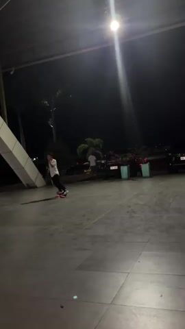 A person is skateboarding at night. The person is wearing a white t-shirt and black pants. The skateboard has lights on the wheels that glow red. The person is covering their face with their hands. The background is dark and appears to be an outdoor area with some buildings and trees. A bright light is shining down from above.