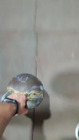 A person's hand, wearing a fitness tracker, holds a metal object with a rounded top and a wider base, appearing to be a kettlebell. The object has a dark, weathered patina with areas of gold and blue. The hand is positioned over a tiled floor. The shadow of the kettlebell and the hand is cast on the floor, creating an elongated shape with a distinct handle form. The background is slightly out of focus, showing more of the tiled floor and a distant, blurry view of what might be an indoor space with some structures visible. The video cuts between static shots and slightly shaky, moving footage.