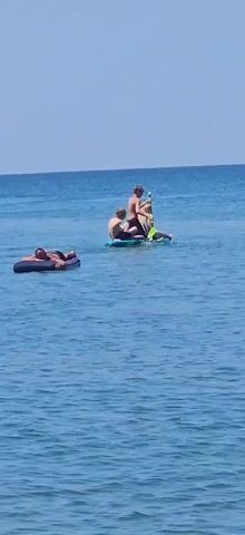 A person lies on an inflatable mattress in the ocean. Further away, two people are on a paddleboard. One person is sitting, and another is standing on their shoulders, holding a paddle. The paddleboard is moving to the right. The sky is clear and blue, and the ocean is calm and blue.