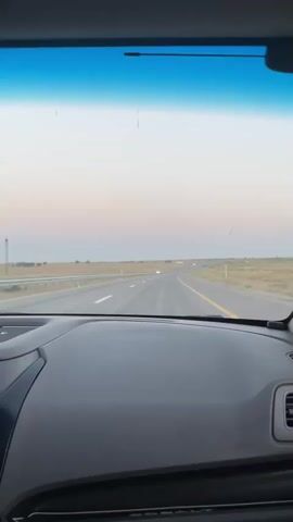 A dash cam records a view from inside a car driving on a highway. The dashboard is visible in the foreground. The highway stretches into the distance, with a beige landscape on either side. The sky is a pale blue and pink, suggesting it is either sunrise or sunset. There are a few other cars visible on the road in the distance.