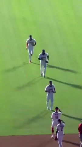 This is an aerial shot of a baseball field, with several baseball players in white uniforms with red accents on the green grass. In the foreground, three players are gathered near the dirt baseline, one of whom is wearing a red shirt. Further up the field, four more players are running towards the foreground. One player in the center foreground is being congratulated by another player. The lighting suggests it's either daytime or early evening with stadium lights on.