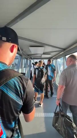 The video shows a group of people walking through an airport jet bridge towards an airplane. The camera is positioned behind a person wearing a black baseball cap and a black and blue patterned shirt, looking forward. Other people in front of them are also walking in the same direction, most wearing similar black and blue shirts. Some individuals are carrying backpacks or bags. The jet bridge has large glass windows on the right side, offering a view of the tarmac and an airplane with a green and white livery. The sky is clear and blue, suggesting daytime.
