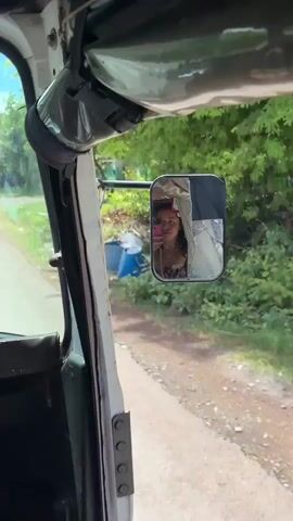 This media file is a video clip taken from the inside of a vehicle, likely a tuk-tuk, looking out through the passenger window and a side mirror. The viewpoint is from the passenger seat, with the dashboard and part of the vehicle's interior visible in the foreground.

The primary subject is a young woman smiling and taking a selfie in the side mirror of the vehicle. She is wearing a colorful, patterned top and a necklace. Her reflection in the mirror is clear, showing her joyful expression.

The setting appears to be a rural or semi-rural area, possibly in Kampot, Cambodia, as indicated by the location context. Outside the vehicle, lush green trees and foliage are visible, suggesting a warm and possibly tropical environment. The road is unpaved and dusty, with some scattered debris along the side, indicating a developing area. The sky is bright and appears to be daytime, with clear weather conditions.

The activity taking place is the woman capturing a moment of her journey by taking a selfie. The overall mood conveyed is one of happiness and enjoyment of the travel experience. The side mirror itself is somewhat damaged, with a crack visible on its surface, adding a touch of realism to the scene.