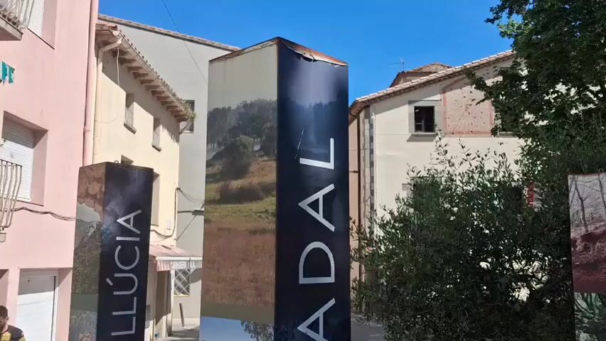 The video shows a street in a small town with black informational pillars. The pillars are marked with white text reading "SANT JULIA - SANTA LLUCIA" on one side and "CANADAL REQUESENS" on another. Each side of the pillars is decorated with a collage of photographs of the town. The buildings are various colors including pink, yellow, and white. The sky is bright blue and there is a tree with green leaves on the side. A car is partially visible parked on the street. The street is paved with gray stones.