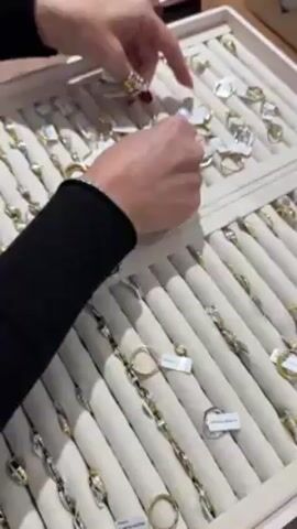 The video shows a jewelry store or display case containing numerous rings and earrings. A person with a black long-sleeved shirt and a bracelet is selecting and handling jewelry. They are pointing to and rearranging the rings, which are mostly gold and silver. The jewelry is neatly arranged in a cream-colored display tray with individual slots for each item, many with small tags.