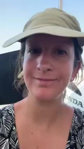 A woman is holding the brim of her hat down so it covers her eyes. She is wearing a black and white print top and a khaki-colored hat. A Honda logo is visible in the background. The sky is blue.