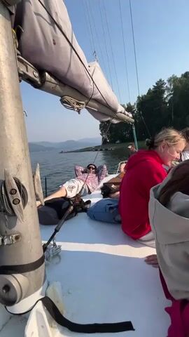 The video pans across the deck of a sailboat on a sunny day. Several young people are visible. In the foreground, on the right, a person wearing a grey hooded sweatshirt and blue jeans sits with their legs crossed, holding a pink bag. Next to them, a person in a red hooded sweatshirt and jeans also sits on the deck. Further back, a person lies on their back, looking up at the sky, wearing a pink and white patterned shirt and light-colored pants. The sailboat has a white deck and a large sail is visible in the upper portion of the frame. In the background, a body of water with a distant treeline and hills under a clear blue sky can be seen. A few small structures or boats are visible on the water in the distance. The camera moves from right to left, then pans slightly to show more of the environment.