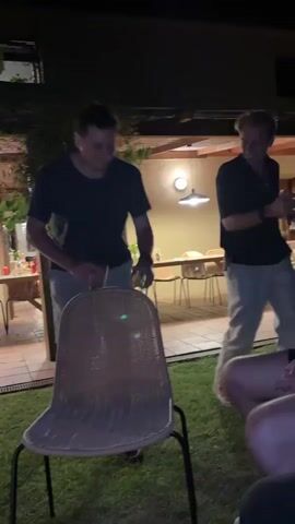 Two men are dancing outdoors at night. The man on the left is wearing a black t-shirt and light-colored shorts. He is holding a clear cup in his right hand and appears to be drinking from it. He is looking to his left and laughing. The man on the right is wearing a black collared shirt and light-colored pants. He is holding a phone in his left hand and looking to his right while dancing. In the foreground, there is a wicker chair with a black metal frame. Behind the men, there is a patio with tables and chairs, and a building with lights on. The ground is covered in grass and paving stones. The overall setting appears to be a backyard gathering or party.