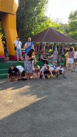 The video shows a group of children in a low crouch on an asphalt surface, as if at the start of a race. They are facing a line of adults standing on a green platform. One adult, a man with a beard wearing a blue sash, appears to be addressing the children. Behind the adults, there are market stalls with colorful fabrics and goods. To the left, a large yellow structure, possibly part of a playground or a stage prop, is visible. The scene is outdoors, with trees and sunlight suggesting it is daytime.