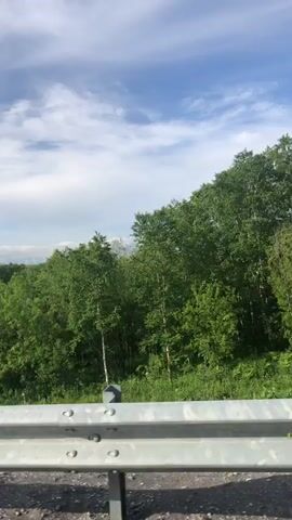 The video shows a view from a vehicle moving along a road, with a large snow-capped mountain in the distance. The foreground is dominated by a metal guardrail. Beyond the guardrail, there is a dense forest of green trees. The sky above is blue with scattered white clouds. The mountain appears to be a volcano, with a cone shape and snow covering its upper slopes and peak. The lighting suggests it is daytime, likely late morning or early afternoon. The overall impression is of a scenic natural landscape viewed from a moving vehicle.