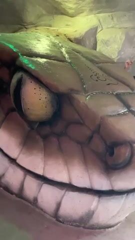 This is a close-up, low-angle shot of a large, sculpted snake head with green lighting illuminating its scales and eye. To the left of the snake's head, a statue of a person in a seated, meditative pose is visible, with their hands pressed together. The background appears to be a cave-like structure with rough, rocky textures and more green lights.