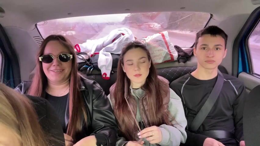 The video features three people riding in the back seat of a car. The woman on the left has long brown hair and sunglasses and is wearing a black leather jacket. The woman in the middle has long brown hair and glasses perched on top of her head. She is wearing a gray sweatshirt and brown vest. The man on the right has short dark hair and is wearing a black long-sleeved shirt with a seatbelt. Behind them in the back of the car are miscellaneous items, including a white jacket and a white plastic bag with red lettering. The camera is facing forward, capturing a view of the interior of the car.