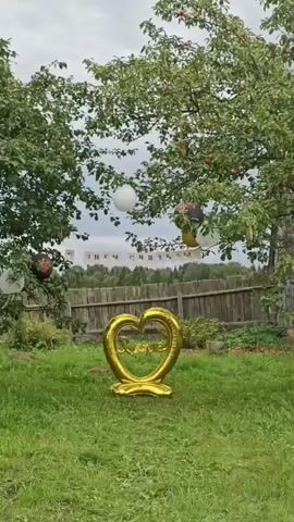 A yard is decorated for an event. In the foreground, a gold heart-shaped balloon that spells "Love" stands on the grass. In the background, several balloons are tied to trees and a banner hangs between two trees. The banner is white with a repeating pattern of flags, with text that is mostly obscured but appears to say "СЧАСТЬЕ" which translates to "happiness." The yard is covered in grass and surrounded by a wooden fence. Trees with green leaves and some red apples are visible, as are some bushes and distant trees. The sky is overcast.