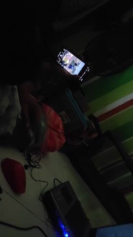 Okay, here is a concise description of the video:

The video appears to be taken from a low angle, possibly from a seated position on the floor in a dim room. The main focus is on a person using a smartphone, seemingly watching video content. The smartphone screen is visible, displaying a video with multiple people. To the right of the person, there is a laptop with the screen lit and an illuminated keyboard with blue lights.  A black mirror or picture frame, a blue storage container, and an orange bag are situated near the laptop. A red, round object, possibly a speaker, sits on the floor next to some tangled cables and black earphones. The background includes a wall with green and white vertical stripes. The overall setting suggests a casual or informal environment, possibly someone's personal space.