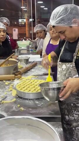 The video shows a cooking class in a kitchen. A person wearing a chef's hat and apron is in the foreground, holding a yellow brush and a metal bowl. They are using the brush to apply something to a pie crust that is in a pie dish. The pie crust has a lattice top. Behind the person in the foreground are several other people, also wearing chef's hats. They are watching the person in the foreground. The kitchen counter is covered in flour and pie crust dough. There are also various kitchen utensils visible, including rolling pins and bowls. The environment appears to be a commercial kitchen or a cooking school.