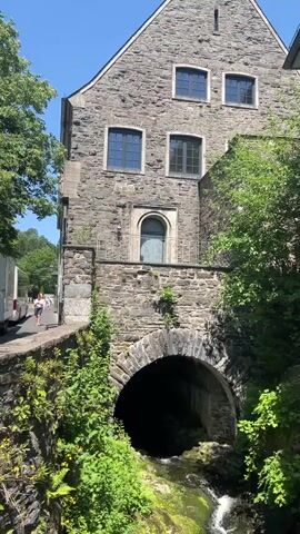 The video shows a stone building with arched windows and a stone bridge over a stream. There are trees and greenery around the building and the stream. A person is walking on a path to the left of the building. The stream has a rocky bed and is flowing quickly. The video is shot on a sunny day.