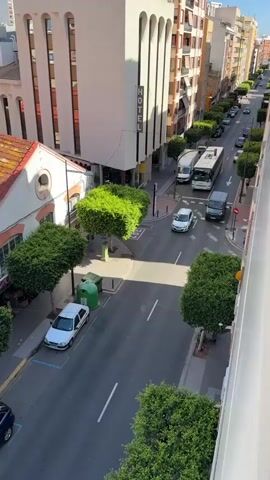This is an overhead, high-angle video shot from a balcony or window overlooking a street in a city. The street is lined with buildings on both sides. The buildings are of various heights and architectural styles, with some appearing to be hotels or apartments. There are trees planted along the sidewalks, which are trimmed into cube shapes. Several cars and a white bus are visible on the street, some in motion and some parked. The sky is clear and blue, indicating a sunny day. The video captures a typical urban scene with traffic and pedestrian activity.