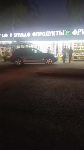 This is a nighttime, eye-level, horizontal video clip showing the exterior of a store at night. The store has a prominent, illuminated sign in Russian. Part of the sign reads "ПРОДУКТЫ" which translates to "PRODUCTS," and "ФРУКТЫ" which translates to "FRUITS," along with a shopping cart icon. To the right of "ФРУКТЫ" is "ШАШЛЫК" which translates to "SHASHLIK" or kebab. The store appears to be a grocery or food establishment, possibly selling fruits and kebabs. In front of the store, there is a display of fruits and vegetables. A dark-colored car is parked to the left of the store entrance. Two men are walking away from the store entrance towards the car, one of them carrying a light-colored bag over his shoulder. The area in front of the store appears to be a sidewalk or paved area, and beyond that is a dark, possibly asphalt, surface that could be a road or parking lot. The lighting is dim, suggesting it is late evening or night, with the store's lights and the car's lights providing illumination. The camera is slightly shaky.
