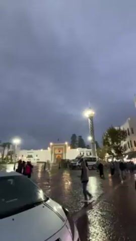 The video shows a wet city square in what appears to be Morocco, possibly Tangier, during twilight or a heavily overcast day. The square is paved with reflective stones, showing standing water.  Several people are walking, some with hooded jackets. A white car is parked in the foreground.  In the background are white buildings, some with architectural details like arches and a tower reminiscent of a minaret.  Streetlights illuminate the scene. There's a structure with a Moroccan flag. At one point, the camera briefly pans to a building with a large coat of arms. Cats are visible near a raised stone structure.