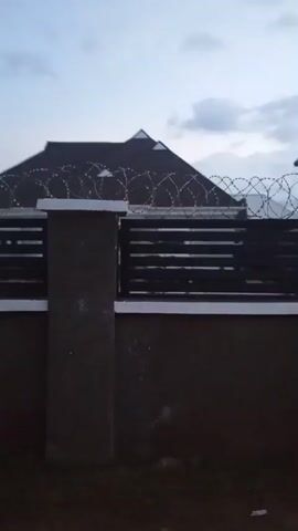 The video shows a dark, cloudy sky with a glimpse of what appears to be a house with a dark roof behind a fence. The fence is made of brown brick with black horizontal slats and a dark grey pillar in the center. Above the fence, barbed wire is visible, suggesting a security measure. The camera is panning slowly to the left, revealing a black gate. The lighting suggests it is either early morning or late evening. There are no people clearly visible in the frame.