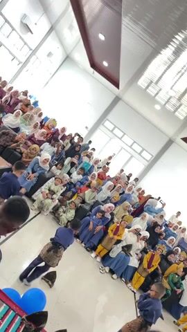 A high-angle shot shows a group of children, dressed in traditional clothing, performing what appears to be a cultural dance for an audience seated on the floor. The audience, comprised of mostly women and children, is dressed in various colorful hijabs and traditional attire. They are seated on the floor of a large, open room with a high ceiling and large windows that allow natural light to enter. Some audience members are seen holding up their phones, presumably to record the performance. In the foreground, two boys are engaged in the performance, while another boy in a blue shirt is also visible. The scene suggests a festive cultural event or celebration.