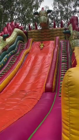 The video shows an inflatable dragon slide with a child sliding down the orange slide. The dragon has green and red scales and yellow accents. The slide is surrounded by other inflatable structures. The child is wearing a yellow shirt and shorts. The video is shot from a slightly upward angle.