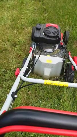 A person is mowing a lawn with a red and black lawnmower. The lawn is overgrown with grass and some yellow flowers. There is a small white building in the background, possibly a shed or cabin, with trees and foliage surrounding it. The camera angle is from the perspective of the person mowing, looking down at the lawnmower and the grass being cut. The footage appears to be from a video recording, as indicated by the timestamp in the corner.