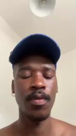 A man is recording a close-up video of himself. The man is light-skinned with short, dark hair and a mustache. He is wearing a blue baseball cap and a small gold hoop earring on his left ear. He appears to be shirtless. The background is a white wall with a white ceiling light. The man is looking at the camera and speaking.