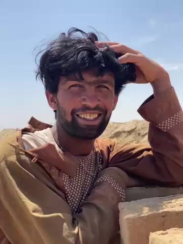 The video features a close-up portrait of a man in an arid outdoor setting. He has dark, curly, somewhat disheveled hair and a dark beard. His eyes are dark, and his face shows signs of being weathered by the sun, with tanned skin. He is wearing a brownish-gold long-sleeved tunic, possibly a *shalwar kameez* top, with intricate white patterned embroidery visible on the chest and cuffs, over a white undershirt. A thin necklace is also visible.

Throughout the clip, the man's expression varies; he is seen smiling broadly at times, revealing stained teeth, and then shifting to a more thoughtful or neutral gaze. His right hand is raised to his head, appearing to scratch or adjust his hair. He is leaning against a light-colored, rough-textured structure, possibly made of mud bricks or similar local building materials.

The background reveals a clear, bright blue sky, indicating a sunny day. Behind him, the landscape consists of sandy or dusty hills, characteristic of a desert or arid region, consistent with the Deh-E Bagh, Afghanistan, location. The strong sunlight casts soft shadows, highlighting the contours of his face and the texture of his clothing. He appears to be engaged in conversation, as indicated by his changing expressions and mouth movements.