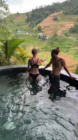 A full shot shows a man and a woman relaxing in a hot tub outdoors, overlooking a scenic valley. The woman is on the left, facing away from the camera, wearing a black bikini. The man is on the right, facing towards her, wearing swim trunks. Both are in the water, which appears to be bubbling slightly, indicative of a hot tub or jacuzzi. In the background, there is a vast mountainous landscape with terraced fields, some green and some light brown, suggesting cultivation. Small houses are visible scattered across the valley. The sky is partly cloudy, and lush green foliage, including palm fronds, frames the scene on the left, adding to the tropical or resort-like ambiance.