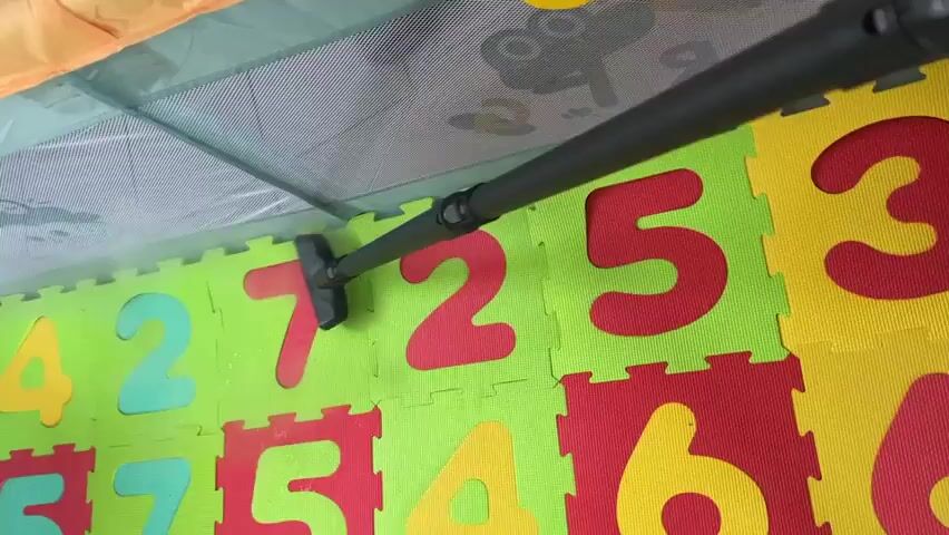 A black vacuum cleaner hose with a black nozzle is seen from above, cleaning a colorful floor made of puzzle mats with numbers. The mats are green, yellow, and red, with large numbers in red and yellow. The vacuum nozzle is positioned over the mats, appearing to be sucking up dust or debris. A portion of a playpen, with a green mesh side and a yellow toy hanging from it, is visible at the top of the frame. The floor appears to be inside the playpen. The lighting is bright, suggesting an indoor environment.